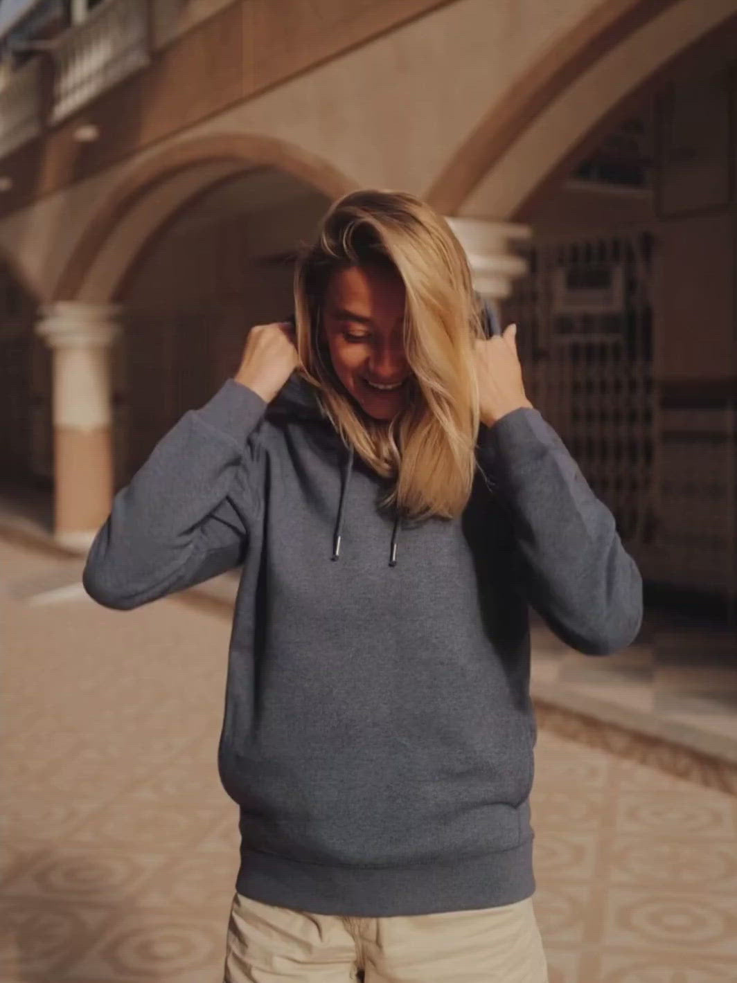 Side dámská mikina s kapucí a kapsami z biobavlny modrá žíhaná žena si vyndá ruce z kapsy a uměje se