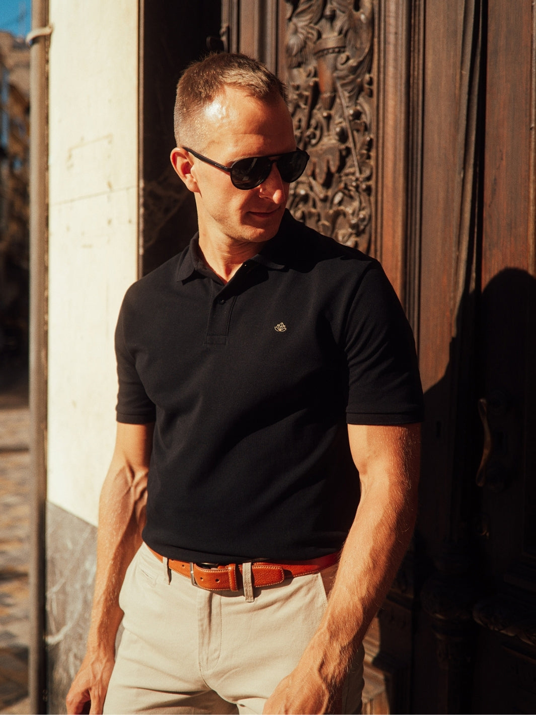 Iconic pánské polo triko z biobavlny černé muž stojí u dřevěných hnědých dveří