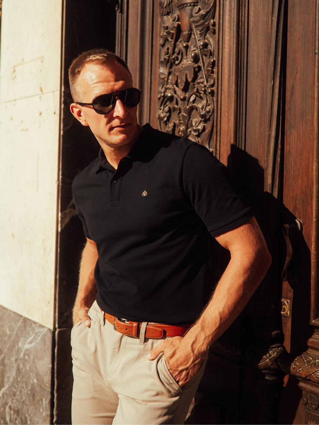 Iconic pánské polo triko z biobavlny černé muž se obírá o dřevěné hnědé dveře